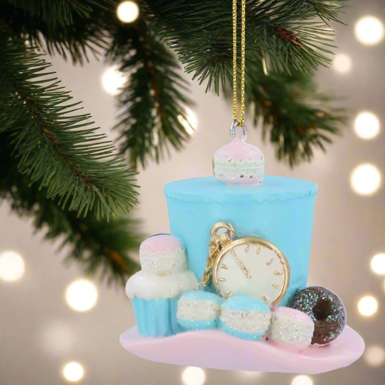 A decorative Christmas tree ornament featuring a teal hat with a golden clock, white icing, and pink sprinkles, set against a blurred background of lights.
