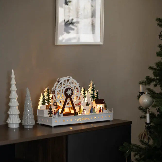 A decorative wooden ferris wheel Christmas scene with LED lights, placed on a table next to a Christmas tree and other holiday decorations.