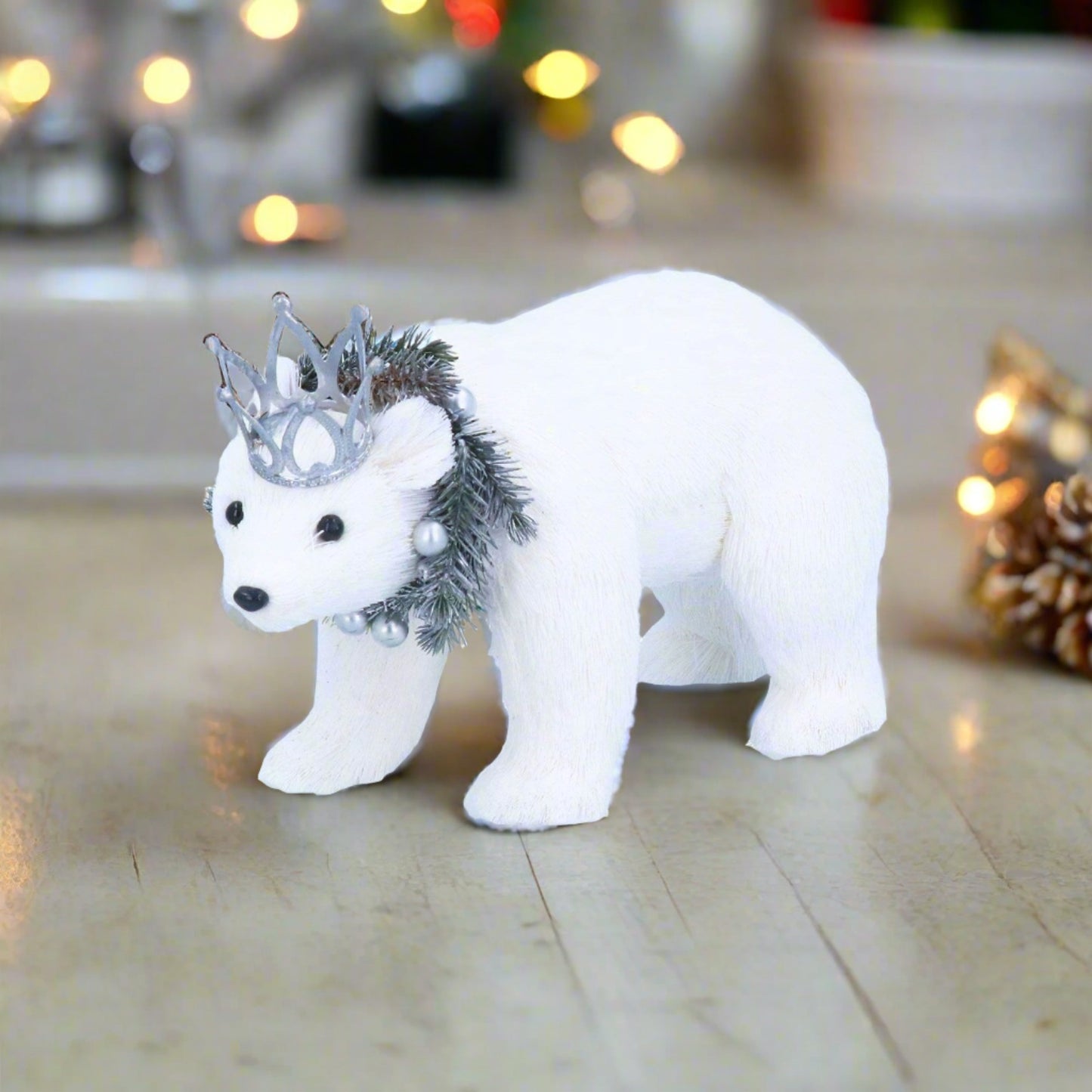 A white polar bear with a silver crown and wreath on a table, with blurred Christmas lights in the background.