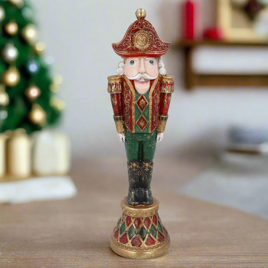 A vintage-style nutcracker Christmas ornament dressed in an elegant, antique-looking uniform with a mustache, positioned on a wooden surface with a blurred Christmas tree and gifts in the background.