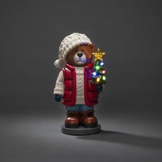 A decorative teddy bear wearing a knitted hat and a red and white coat, holding a Christmas tree with multicolored LED lights.