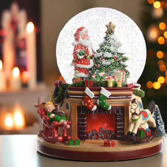 A Christmas-themed snowglobe featuring a fireplace scene with Santa Claus, a Christmas tree, and holiday gifts inside the globe. The base is brown, and there is a blurred warm light background.