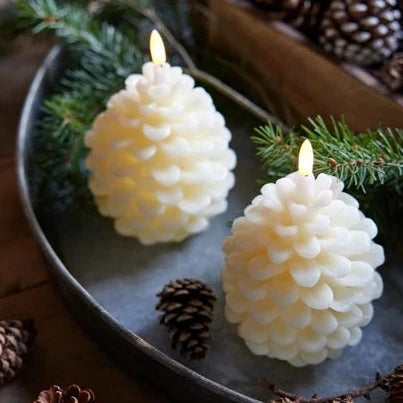 Two cream-colored pinecone-shaped LED candles placed in a metal dish with pine branches and decorative pine cones around.