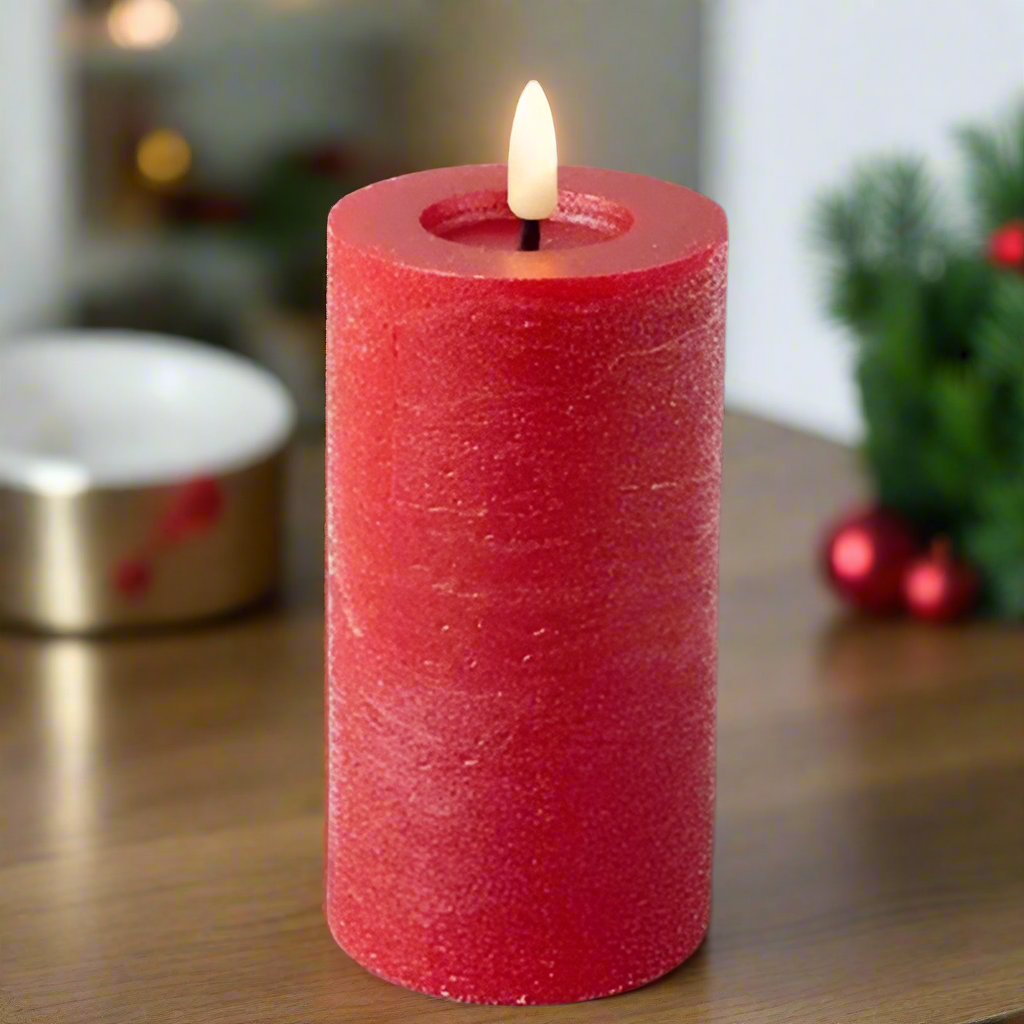 A red, real wax candle with a lit flame, placed on a wooden surface, with Christmas decorations in the background.