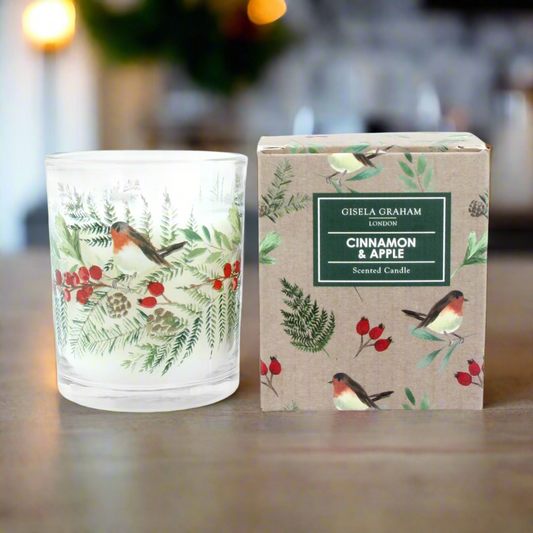 A clear glass scented candle with a Christmas design, featuring a robin, cinnamon, and apple, alongside a cardboard box with a matching design.