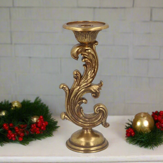 A gold-colored single arm candlestick holder with a scroll design, placed on a white surface with Christmas decorations around it.
