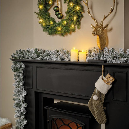 Pre-lit Christmas garland with green foliage and snow effect, featuring warm white LED lights, displayed over a fireplace with a reindeer decoration and a stocking hanging below.