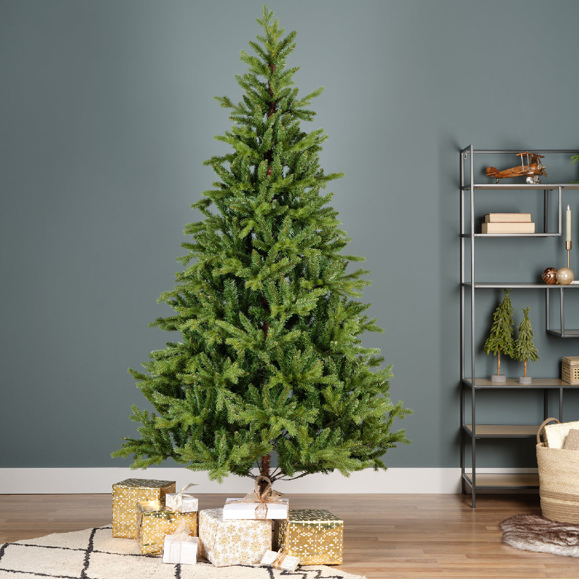 A realistic green pine Christmas tree with metal feet, placed on a decorative mat, surrounded by holiday decorations and books in the background.