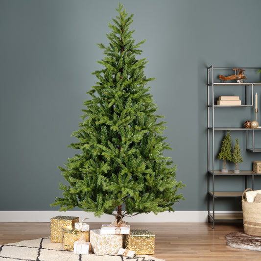 A realistic green pine Christmas tree with metal feet, placed on a decorative mat, surrounded by holiday decorations and books in the background.