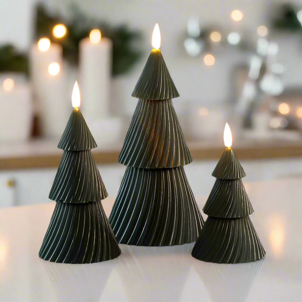 Three green Christmas tree wax candles with realistic flickering effect, placed on a white surface in a home environment with candles in the background.