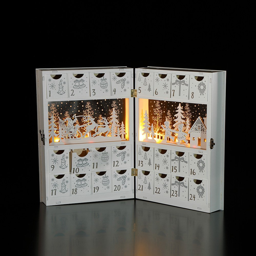 A white wooden book advent calendar with LED lights illuminating a snow scene cut out, opened to reveal numbered drawers.