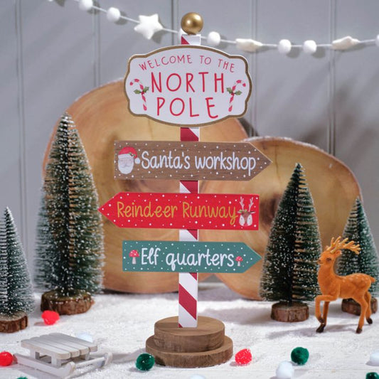 A wooden sign post with Christmas decorations, featuring signs that say 'Welcome to the North Pole', 'Santa's workshop', 'Reindeer runway', and 'Elf quarters'. There are miniature trees and a reindeer figure in the background.