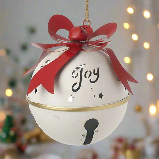 A white Christmas bell with the word 'joy' cut out, accented with a red bow and jute rope, hanging in a festive setting with Christmas lights.