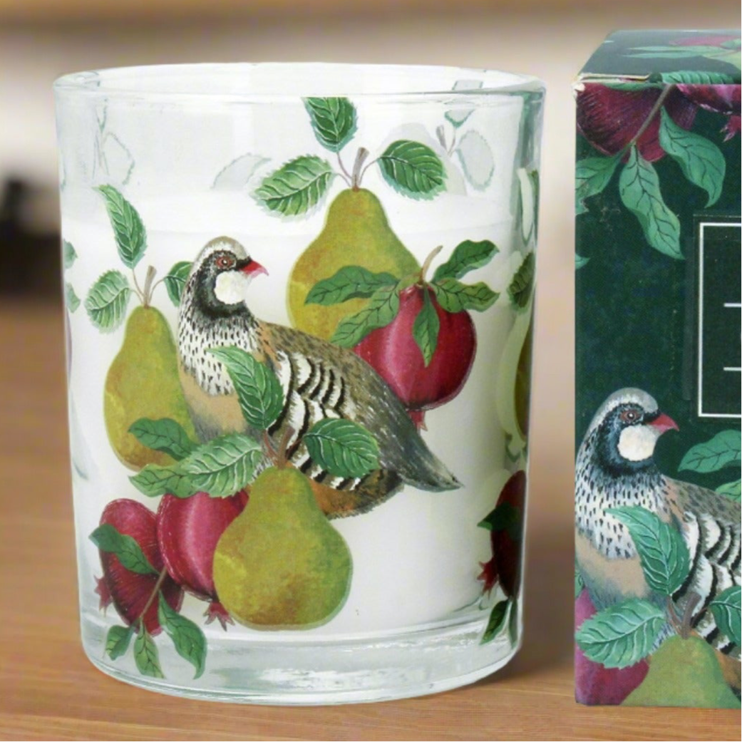 A cylindrical glass candle with a partridge and pear design, sitting next to its packaging box.