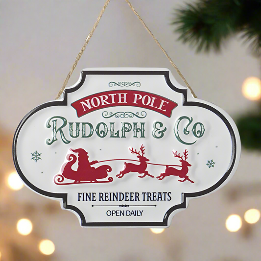 A circular wooden sign with a Christmas theme, featuring the text 'NORTH POLE RUDOLPH & CO FINE REINDEER TREATS', accompanied by images of Santa Claus and a reindeer. The sign is hanging from a metal jute rope.