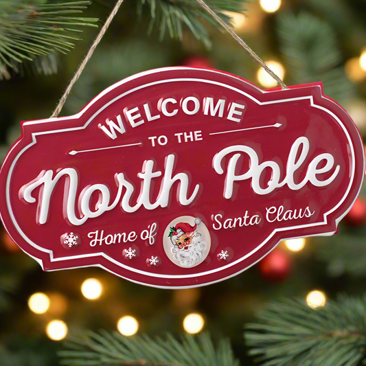 A red and white 'Welcome to the North Pole' sign with a Santa Claus design, hanging from a Christmas tree.