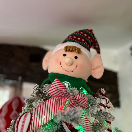 A smiling elf head tree topper placed on a Christmas tree, wearing a red and green hat with a happy facial expression.