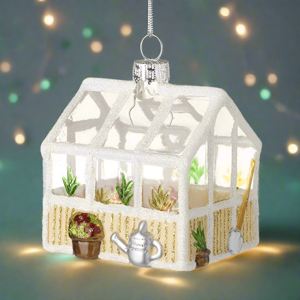 A glass ornament in the shape of a greenhouse with plants and tools inside, hanging from a silver loop.