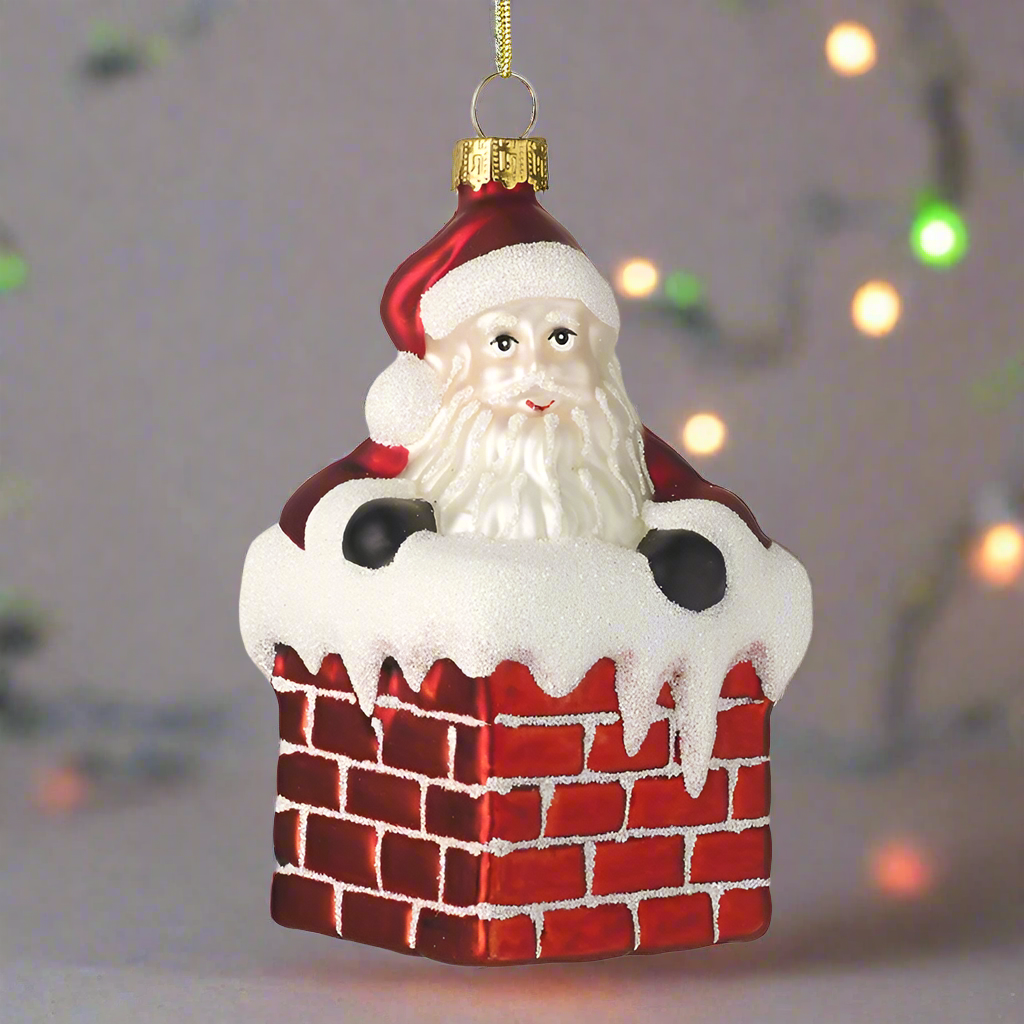 A glass decoration featuring Santa Claus with a red suit and white beard, peeking out from a red chimney with white snow on it, designed for hanging on a Christmas tree.