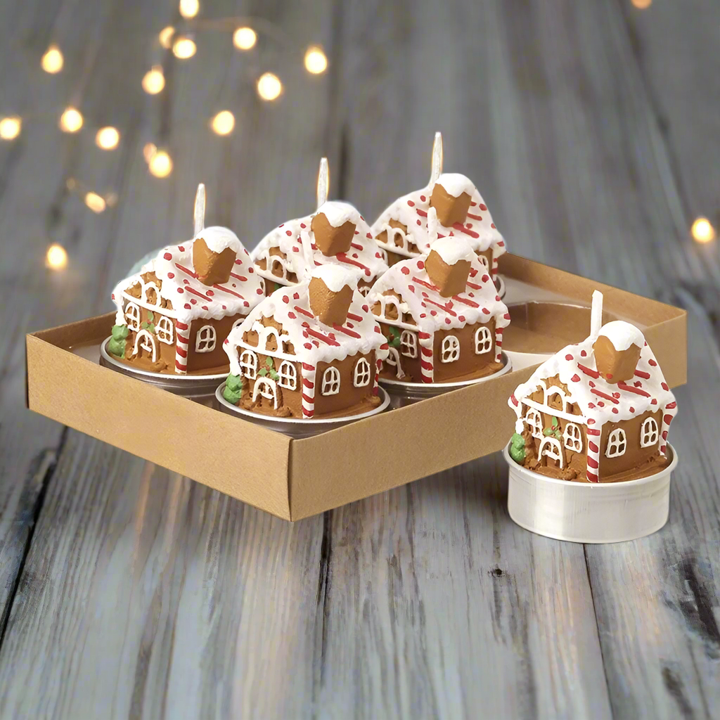 A set of six gingerbread house-shaped tea lights in a cardboard box, with white frosting and red and green candy decorations.