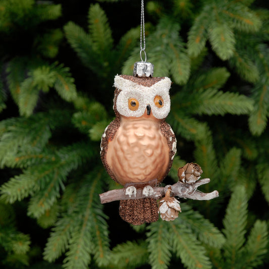 A glass ornament depicting an owl perched on a branch with miniature pine cones, hanging from a silver wire.