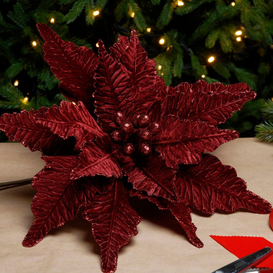 A close-up image of a burgundy velvet Christmas poinsettia stem with artificial berries, placed on a table with a blurred Christmas tree and presents in the background.