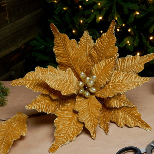 A gold velvet artificial poinsettia stem with pearls, placed against a festive background with Christmas lights.