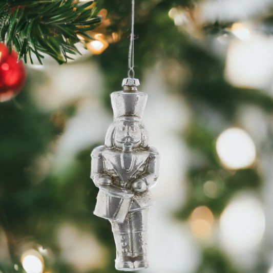 A silver nutcracker hanging from a Christmas tree, dressed in a uniform, with a blurred festive background.