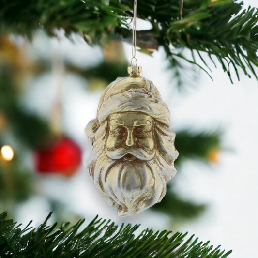 A gold-colored Santa head hanging decoration bauble placed on a Christmas tree with blurred lights and a red ornament.