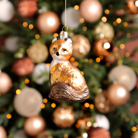 A glass fox hanging Christmas bauble decoration with a golden hanging wire, displayed against a backdrop of round decoration lights.