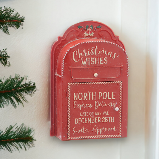 A red vintage-style Christmas wishes North Pole express delivery post box with a white door, hanging on a wall. The post box is decorated with a festive design, and it is placed against a backdrop of a Christmas tree.