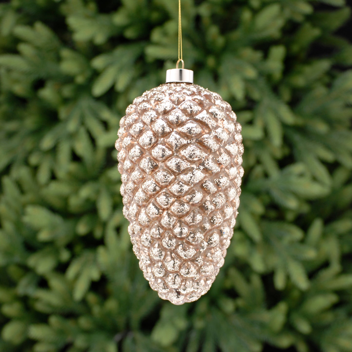 A golden glass pinecone-shaped Christmas decoration hanging against a blurred foliage background.