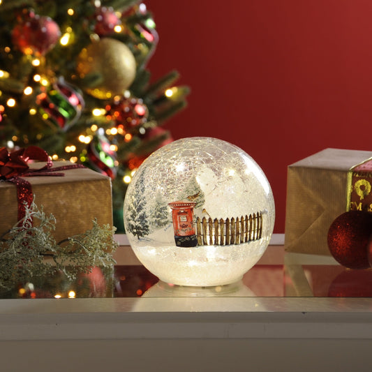 A lit crackle effect Christmas ball with a winter scene post-box, placed on a table with Christmas decorations in the background.