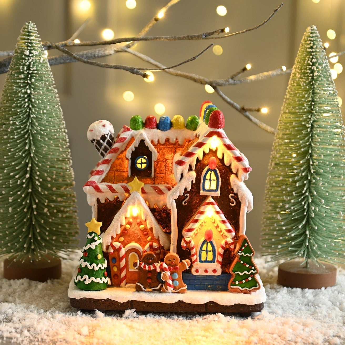 A resin gingerbread house decoration with a white, red, and green color scheme, featuring candy-like details, a warm white LED light, and placed on a snowy surface with Christmas trees in the background.