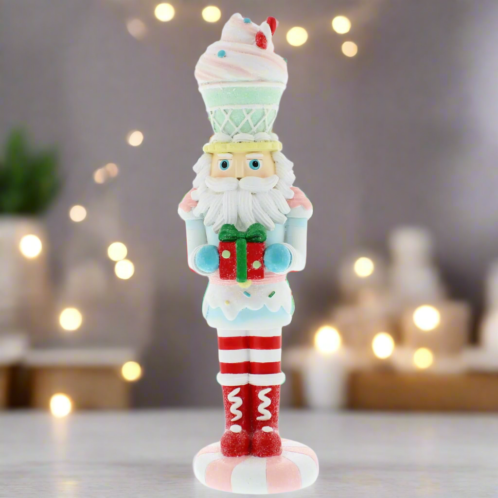 A pastel-colored cupcake nutcracker figurine, with a white and red hat resembling a cupcake, holding a present, standing against a backdrop of fairy lights.