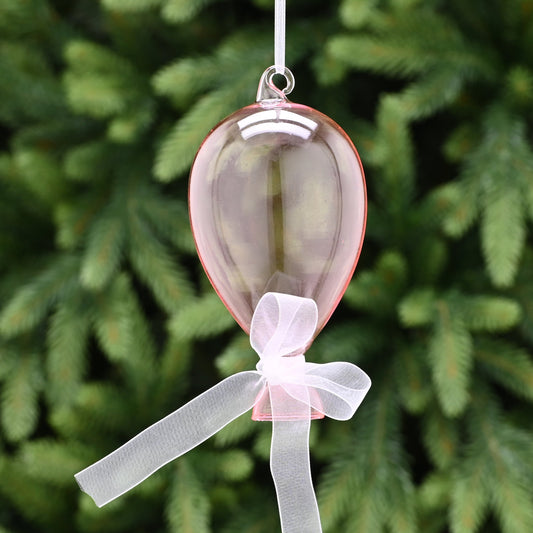 A pink glass balloon with a white ribbon bow, hanging from a tree.