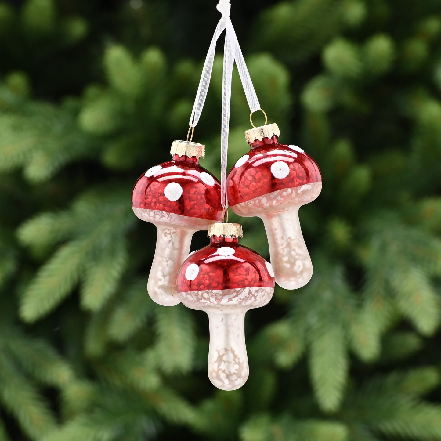 Three red mushroom-shaped Christmas tree decorations with white stems and red caps, hanging from a green tree with a blurred background.