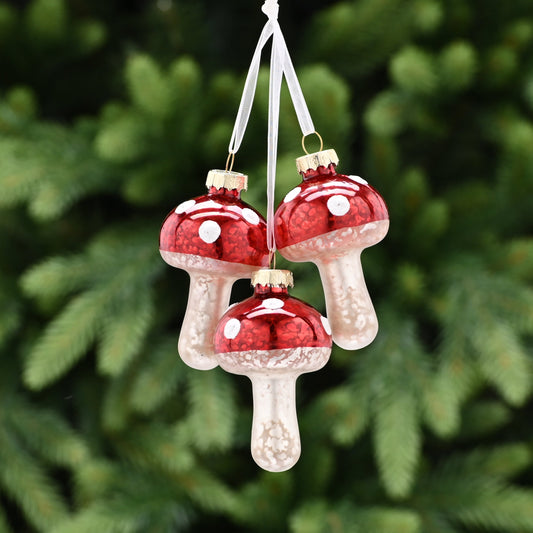 Three red mushroom-shaped Christmas tree decorations with white stems and red caps, hanging from a green tree with a blurred background.