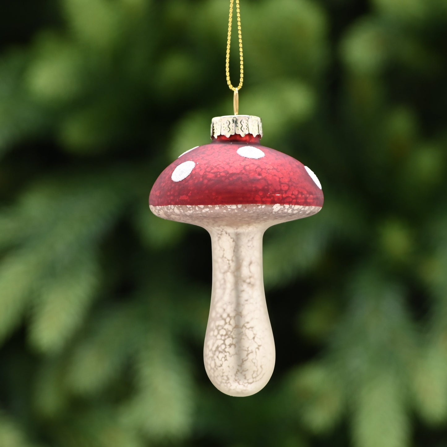 A red and white mushroom-shaped hanging Christmas tree decoration with a gold-colored stem and a tarnished gold interior, set against a blurred greenery background.