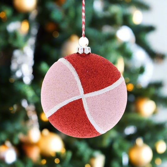 A round pink and red Christmas decoration hanging from a string, resembling a candy cane, displayed against a Christmas tree backdrop.