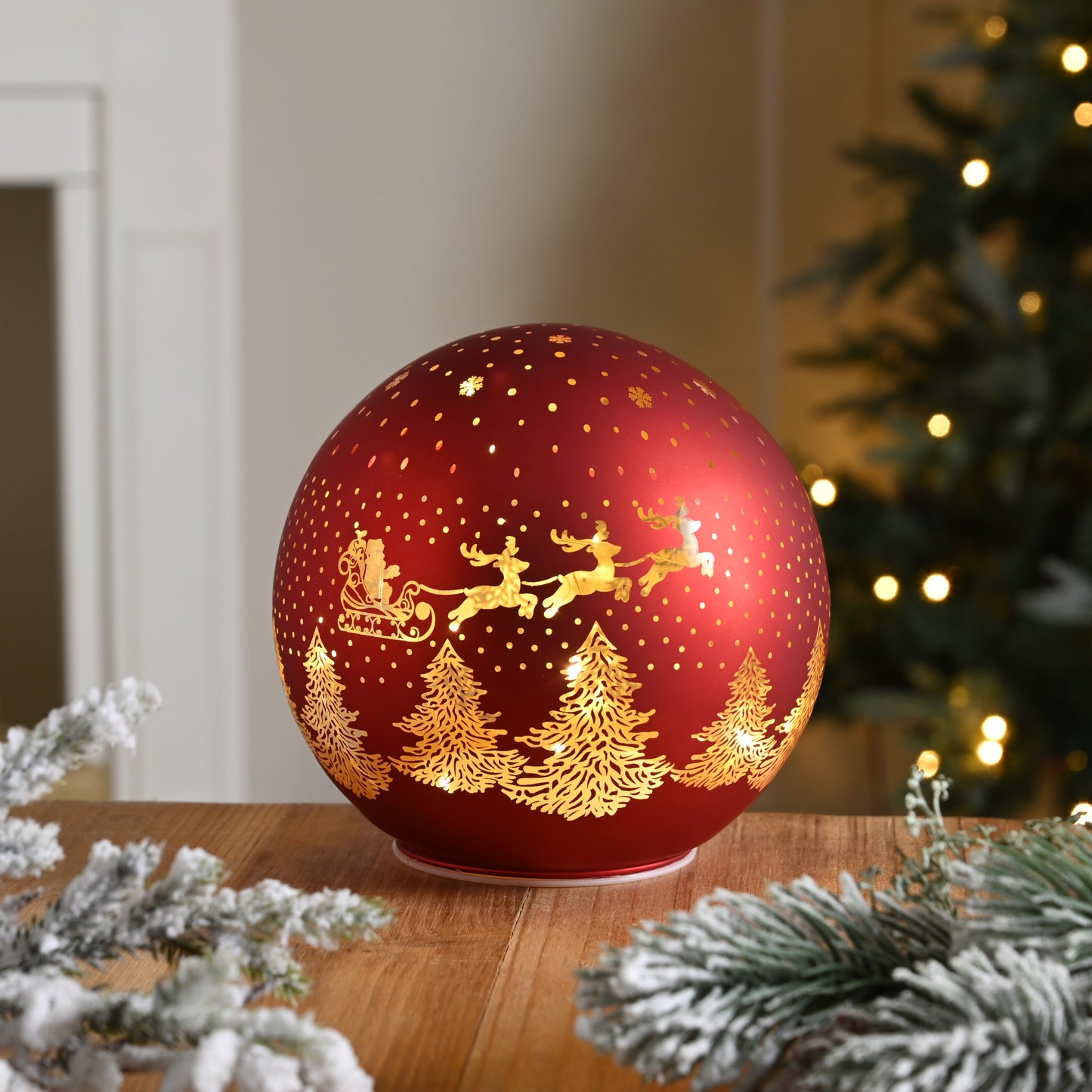 A red light up Christmas decoration featuring a Santa and sleigh scene, placed on a wooden surface with a blurred Christmas tree and lights in the background.