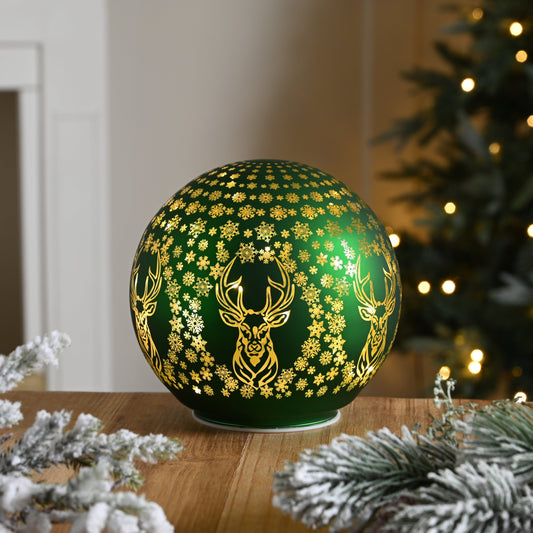 A green light-up ball with a reindeer and snowflake design, sitting on a wooden surface with a blurred Christmas tree and lights in the background.