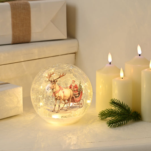 A clear crackle effect ball with a Santa Claus and reindeer figure, illuminated by warm white LED lights, placed on a white surface next to candles and Christmas decorations.