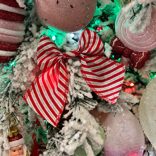 A red and white striped candy cane bow attached to a Christmas tree decoration, with festive lights and other holiday ornaments in the background.