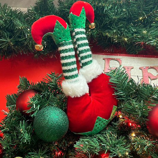 A pair of red elf legs with green accents hanging from a Christmas tree with festive decorations around.