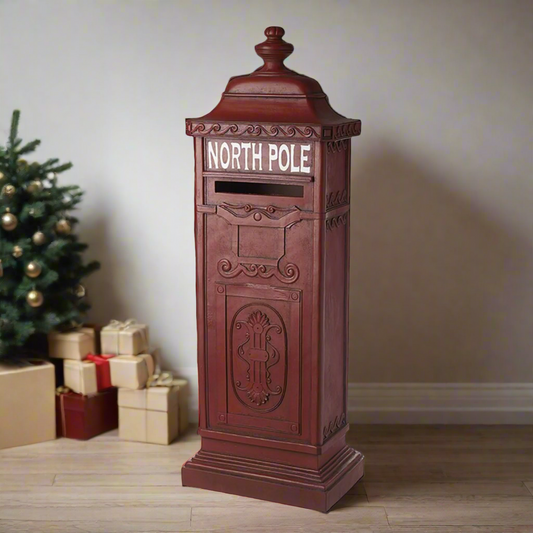 A red vintage style North Pole Christmas post box, positioned indoors with Christmas decorations in the background.