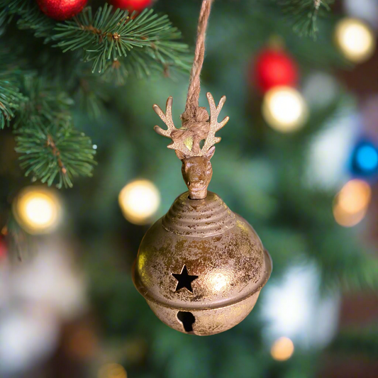 A gold-colored metal sleigh bell with a stag head design, featuring a keyhole in the front, hanging from a Christmas tree with festive lights.