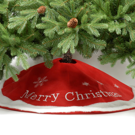 A red Christmas tree skirt with 'Merry Christmas' text and silver embroidered snowflakes, placed under a green Christmas tree with pine cones on its branches.