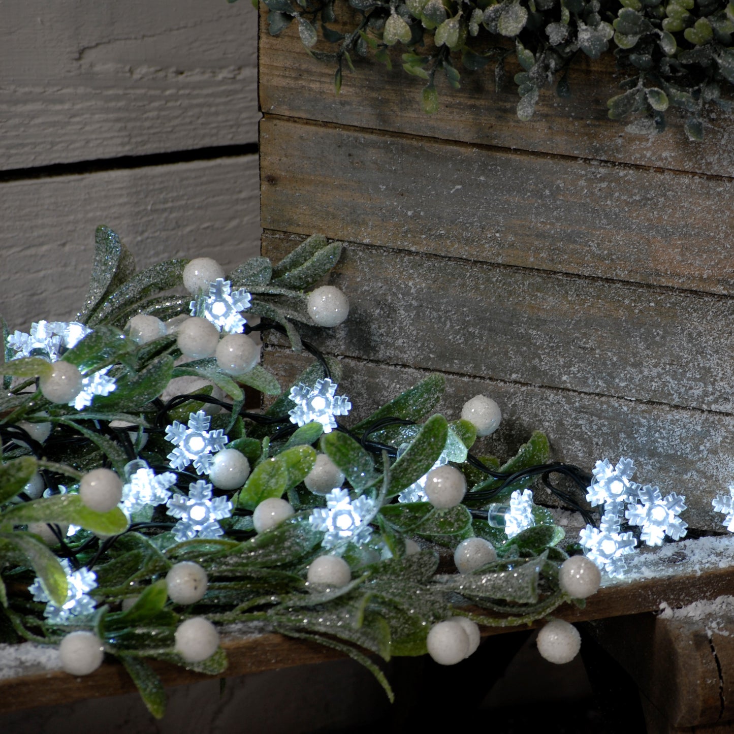 Battery operated white snowflake Christmas string lights with green cable, displayed against a wooden backdrop with artificial greenery.