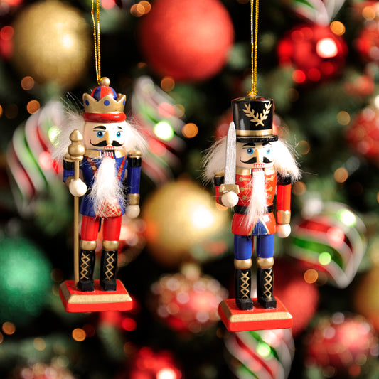 A pair of hanging Christmas nutcrackers decorations, one in blue with a crown and the other in red with a staff, hanging from a tree with Christmas balls and ornaments around.